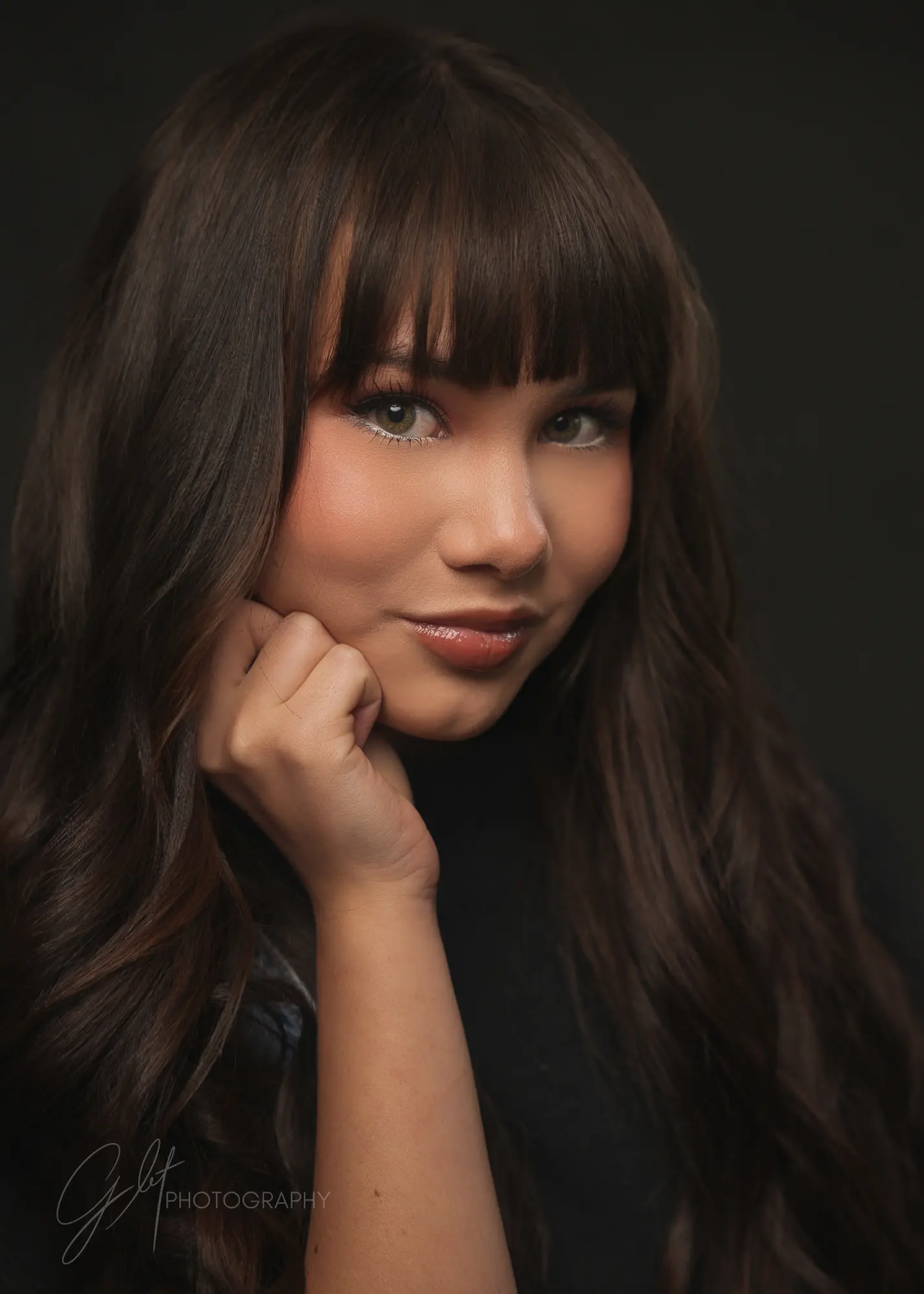 A young woman with bangs, chin resting on her hand, soft studio light